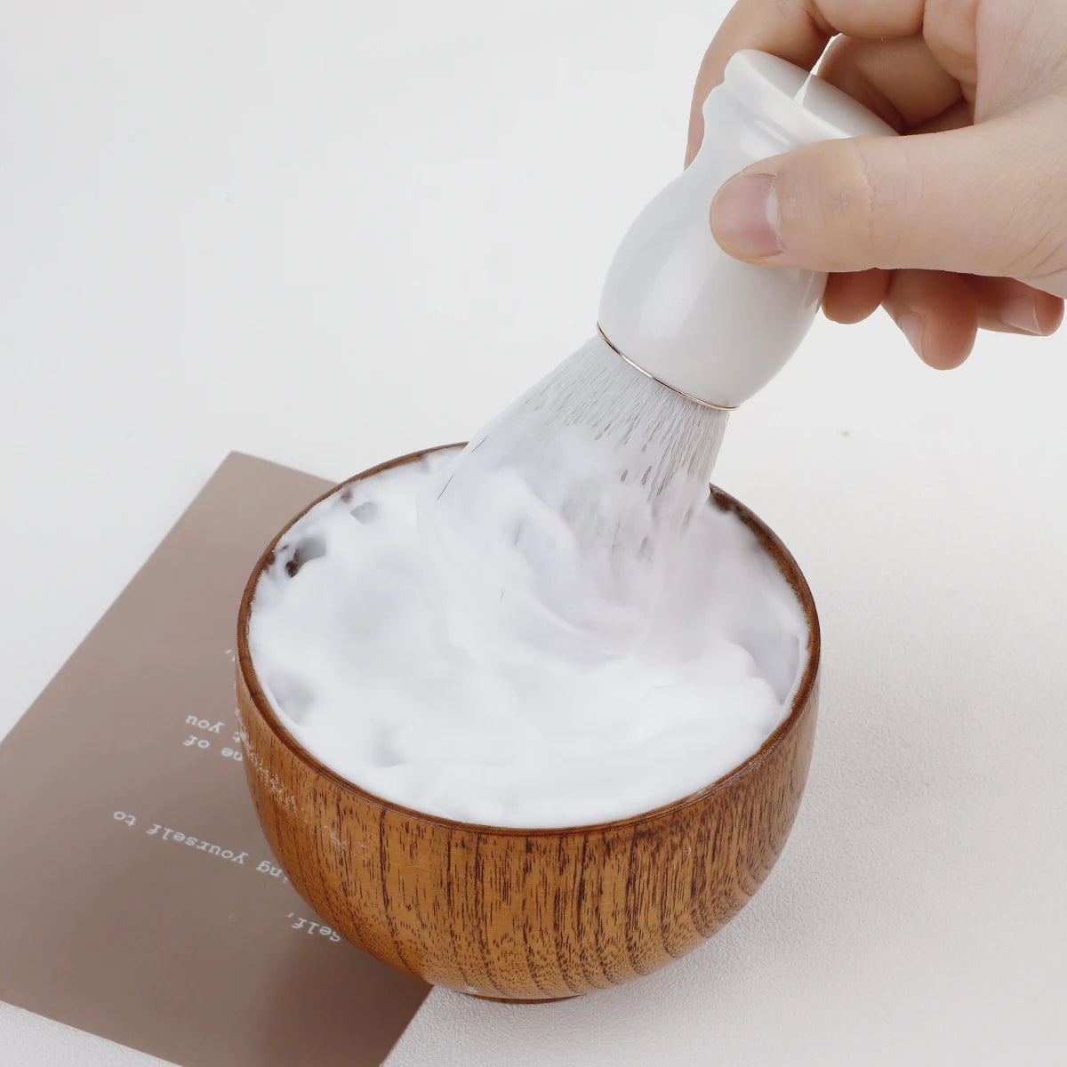 Person using a white brush to scoop foam from a wooden bowl on a light background
