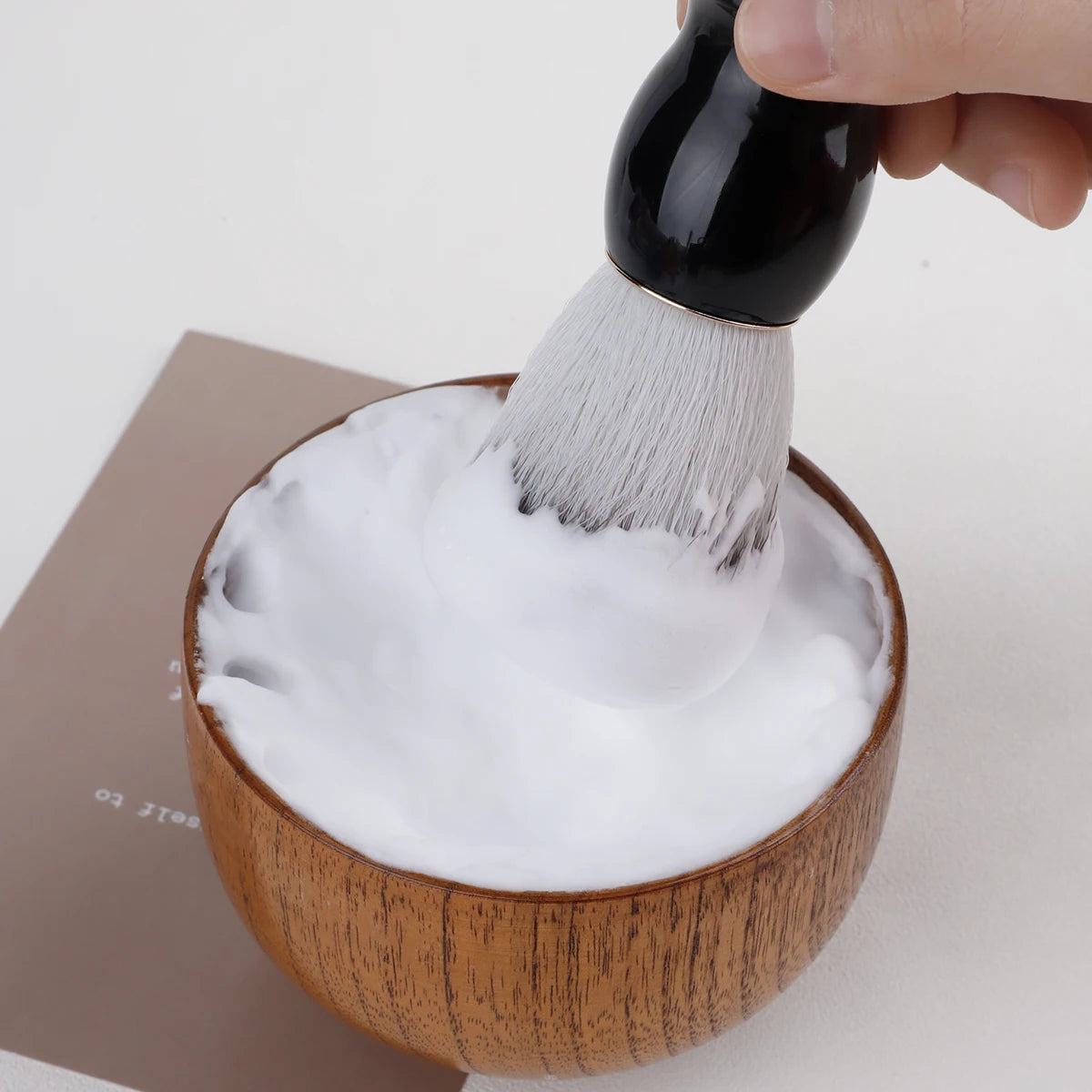 Person using a black brush to scoop foam from a wooden bowl on a white background